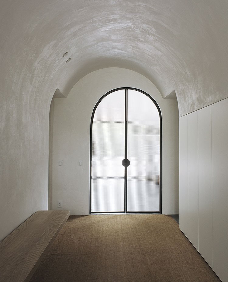 Zona de entrada con banco de madera volado, techo curvo y puerta con cristal esmerilado