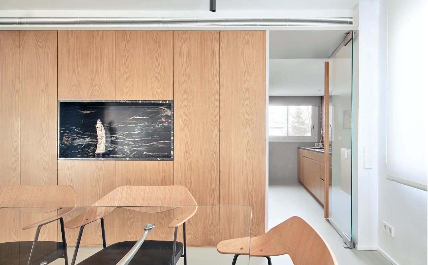 Comedor formal comunicado con cocina, hornacina en frente de almacenaje revestida de mármol, sillas con respaldo de madera, mesa con sobre de cristal