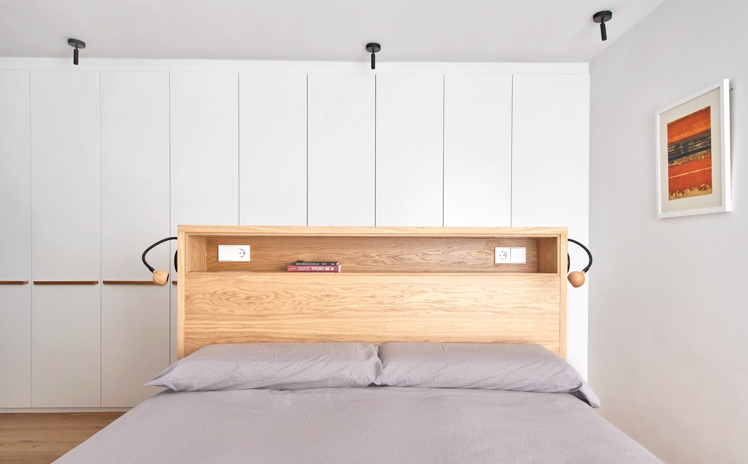 Dormitorio con cama central y amplio frente de armarios de almacenaje en pared en blanco, con tiradores en madera
