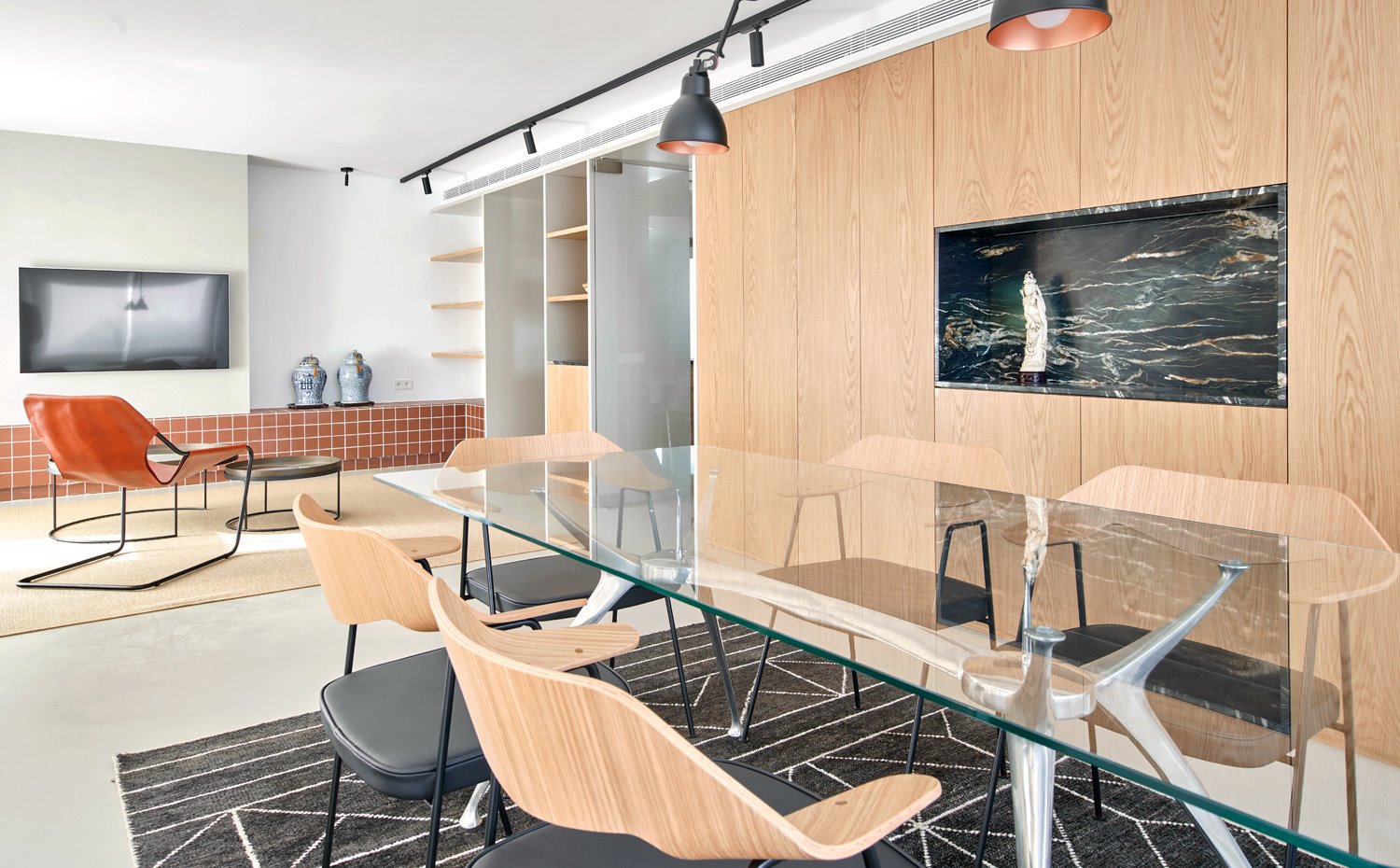 Zona de comedor con sillas en negro y madera, mesa con sobre de cristal sobre al fombra en negro con motivos en blanco, y al fondo butaca de piel y mesitas auxiliares redondas