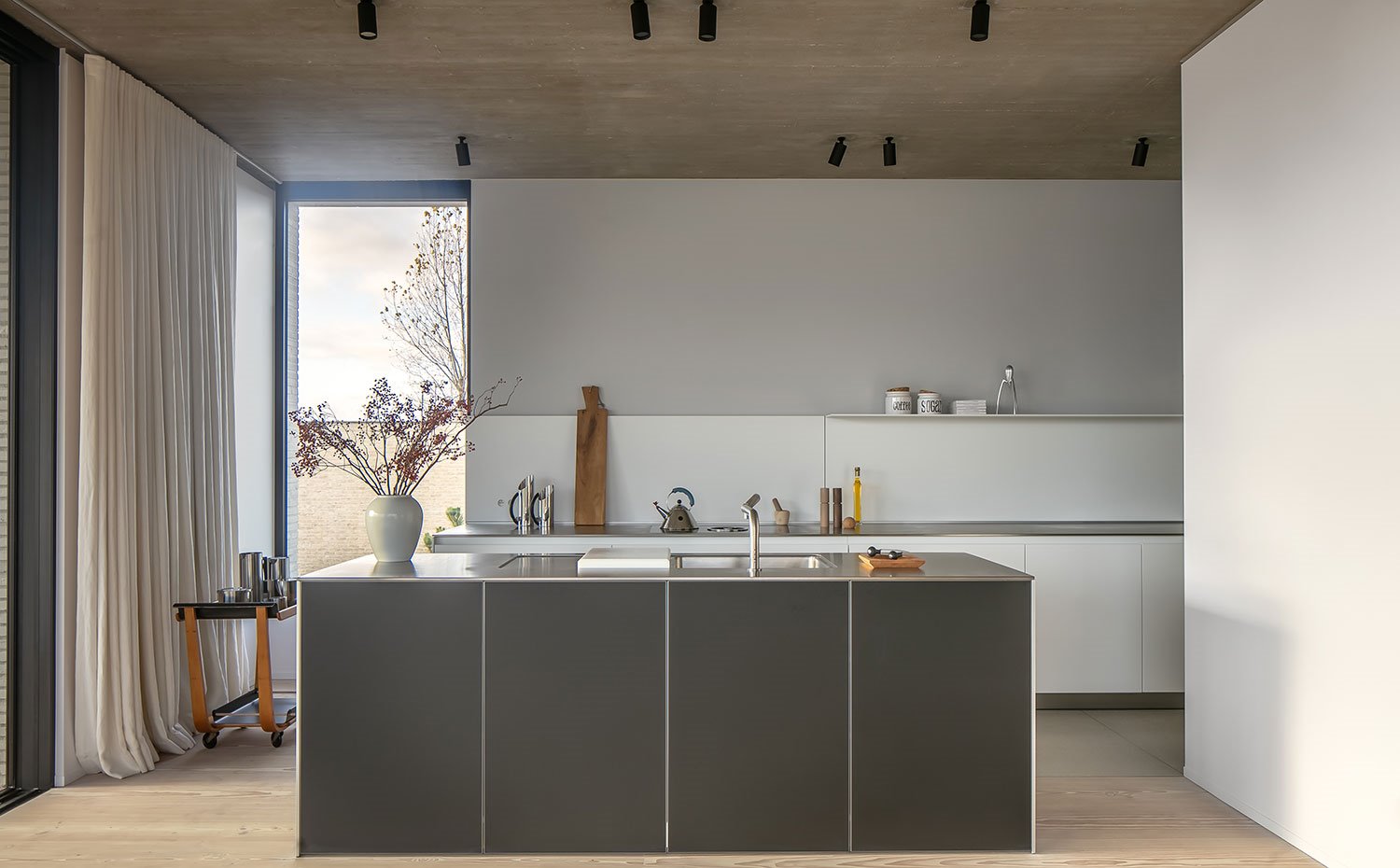 Cocina con isla con frentes en negro, focos techo en negro, suelo de madera, cortinas de techo a suelo en ocre, cerramientos acristalados, carrino de madera