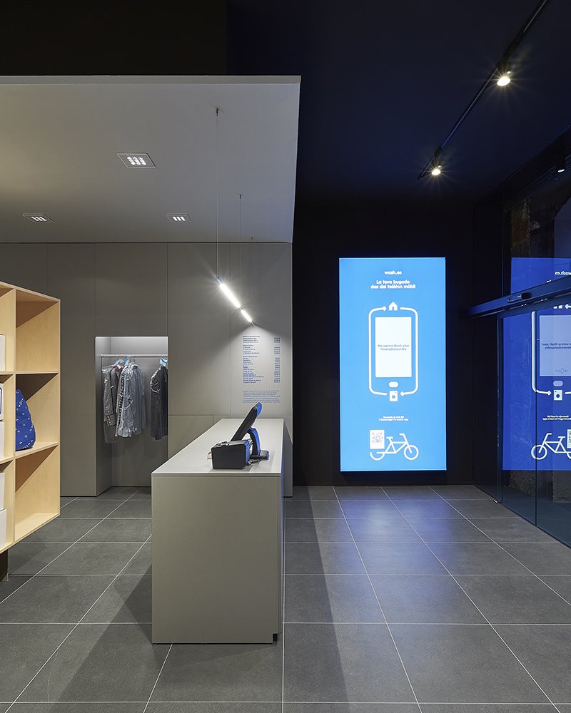 Lavanderia de diseño en Barcelona 4. Como si fuera la pantalla de un móbil gigante, el panel retroiluminado de la zona de entrada recibe al cliente en un baño de luz azul: el color que conecta el mundo virtual de la app con el espacio físico de la tienda.