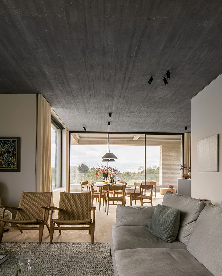Salón con butacas de madera y fibras naturales, sofá en gris, techo de hormigón, al fondo mesa y sillas de comedor