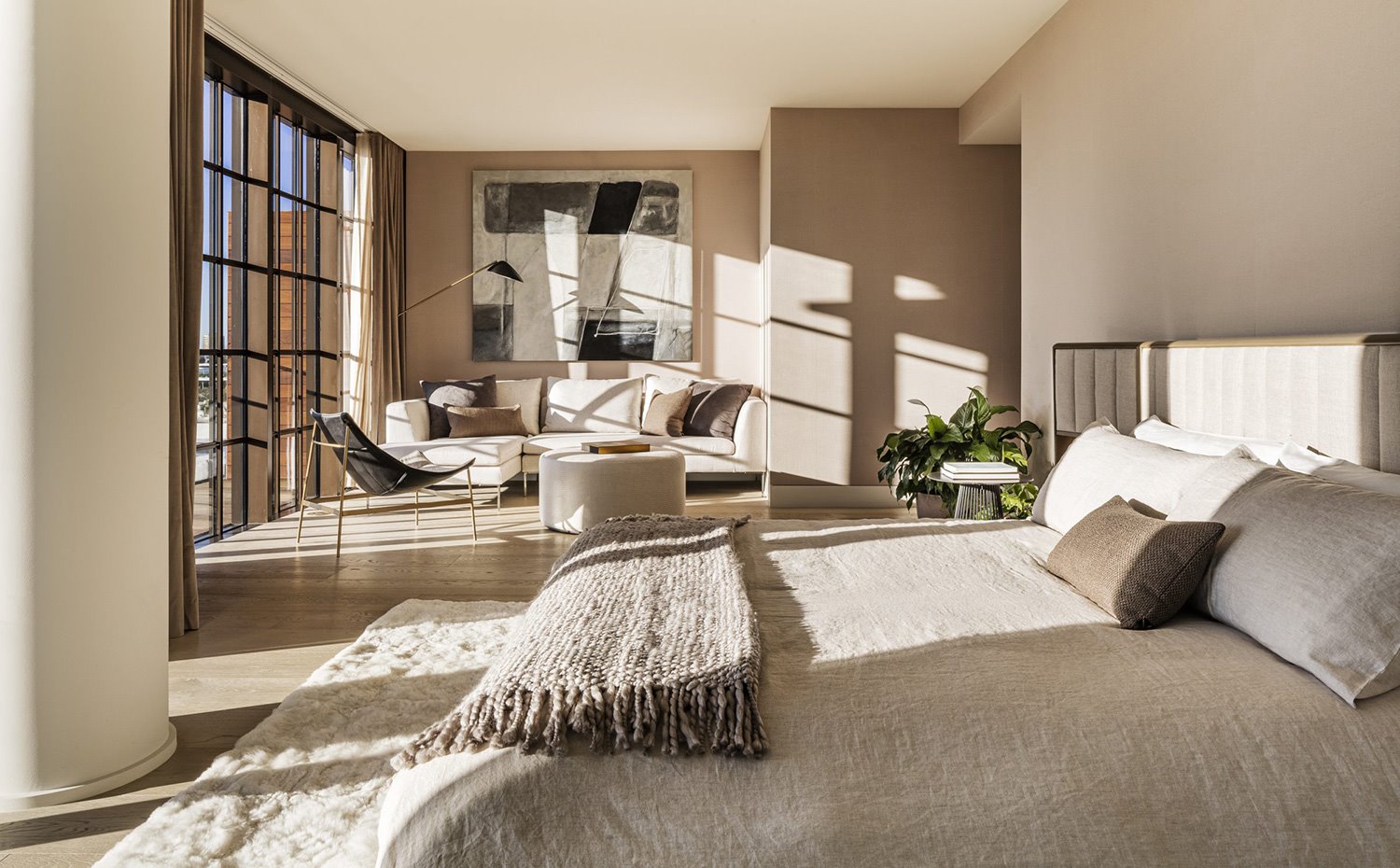 Dormitorio en tonos crudos, con zona de estar con sofá en tonos neturos y butiaca en negro, cerramiento acristalado con perfilería de aluminio