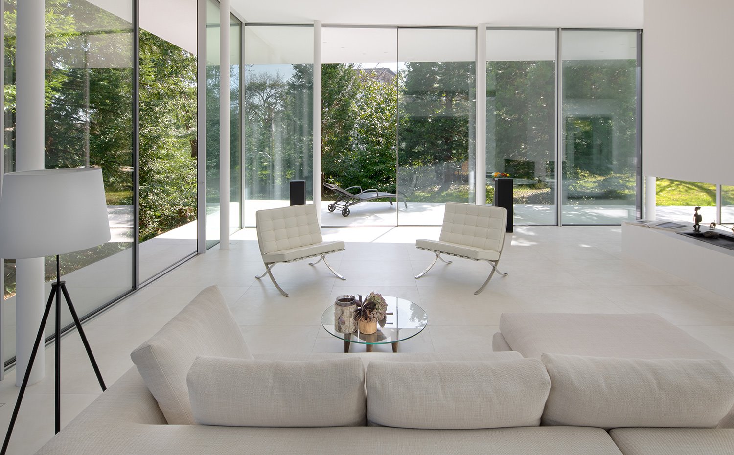 Salón con sofá y dos butacas, en blanco, mesa de centro con sobre de cristal y patas de madera y luminaria de pie, chimenea de obra en el centro de la planta