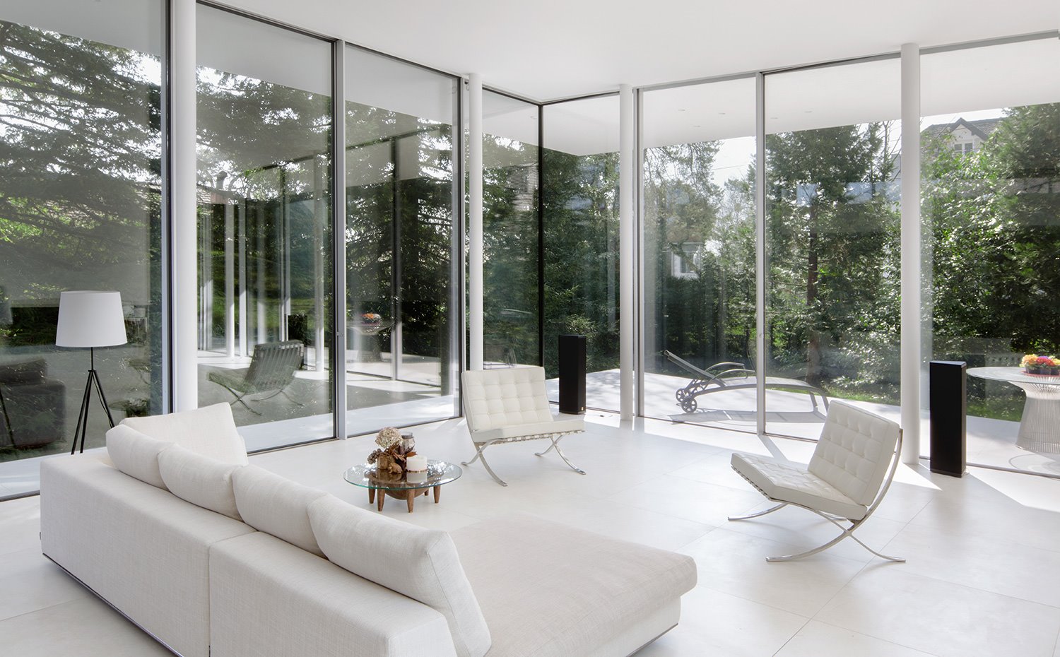 Salón interior con mobiliario en blanco, suelo en blanco, cerramientos en cristal, vistas de los árboles exteriores