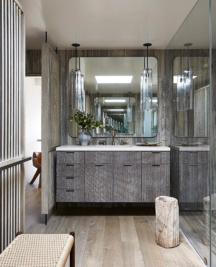 Cuarto de baño con zona de tocador revestida e madera y mármol, espejo cuadrado con cantos curvos, luminarias suspendidas, mampara de ducha en cristal, taburete e madera