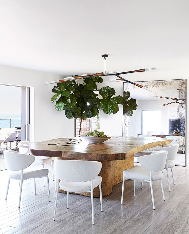 Mesa de comedor de formas irregulares en madera tropical, sillas en blanco, amplias aperturas hacia el exterior