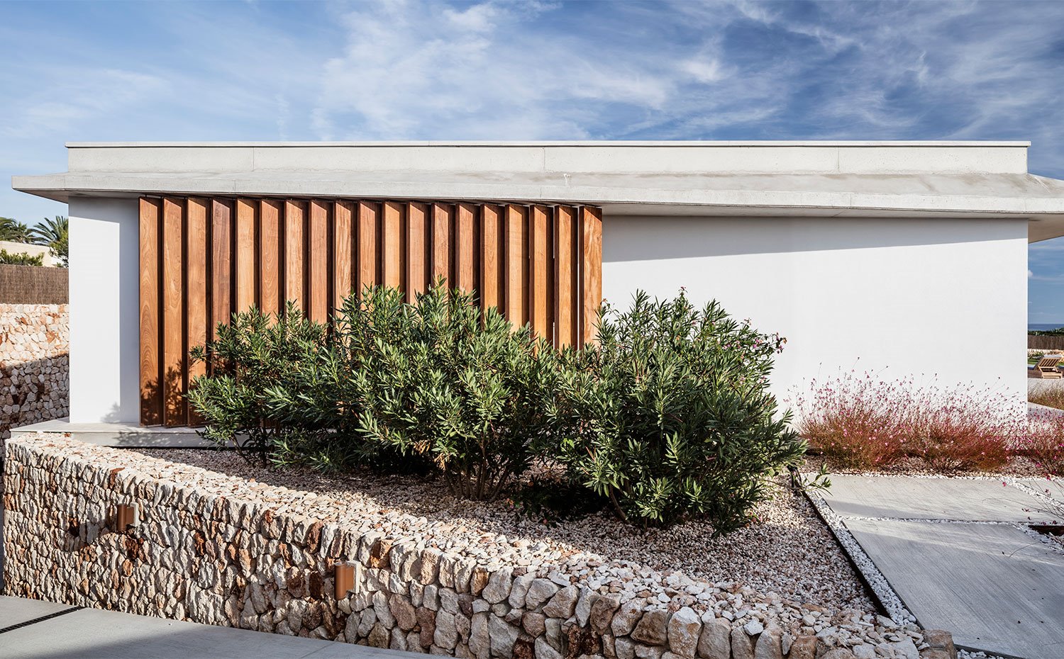 Parte trasera de la vivienda con vegetación sobre piedras de cantos rodadeos y combinación de madera, hormigón y piedra para estructura