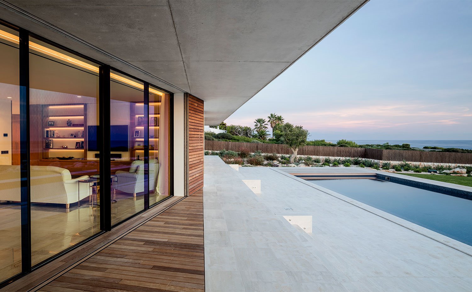 Vista del salón desde el exterior, cubierta de hormigón, suelo exterior de madera y pavimento porcelánica hacia piscina