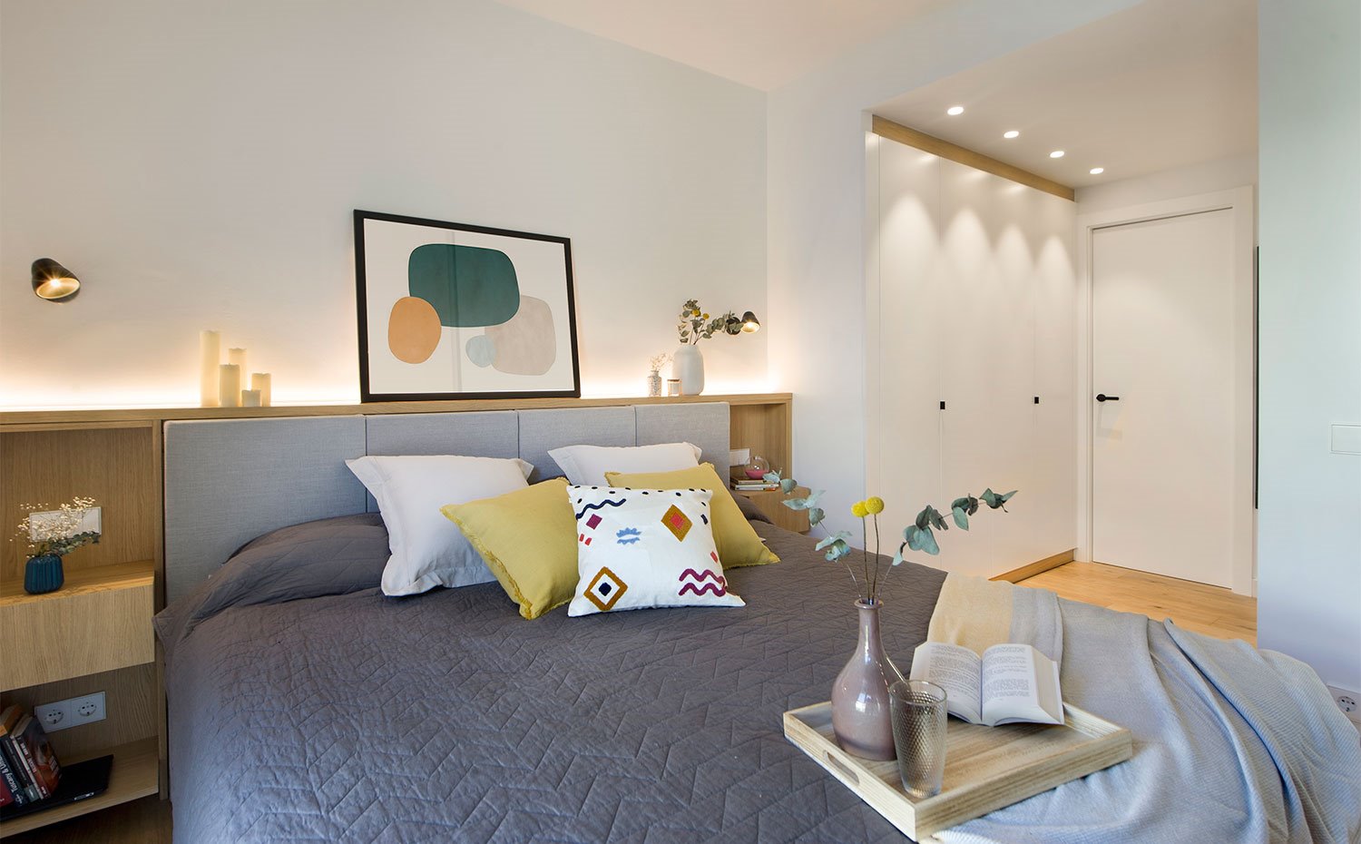 Dormitorio principal con cabecero en madera con tapizado en gris, zona de armarios empotrados en blanco. 