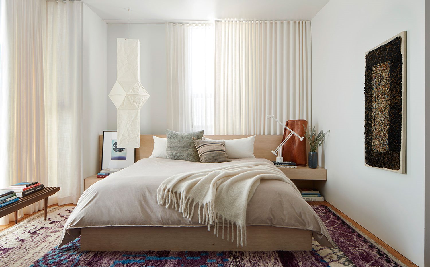 Dormitorio con alfombra multicolor, cortinas en crido, mueble cabecero, bancada de madera con libros, luminaria suspendida de pared