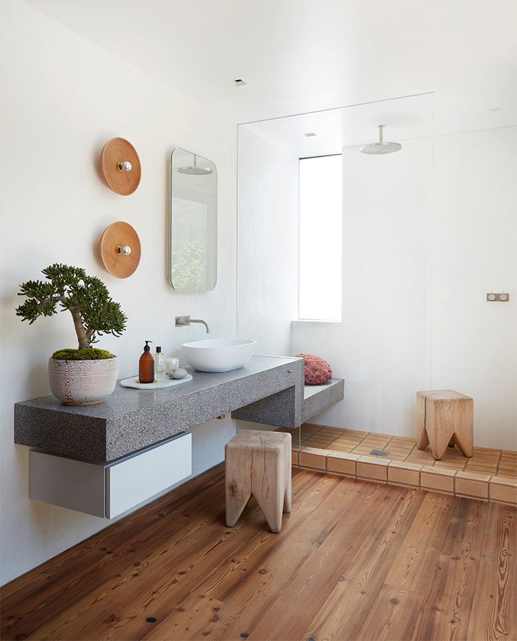 Mueble lavamanos de piedra con extensión de bancada hacia ducha, taburetes de madera, bonsai sobre encimera, suelo de madera