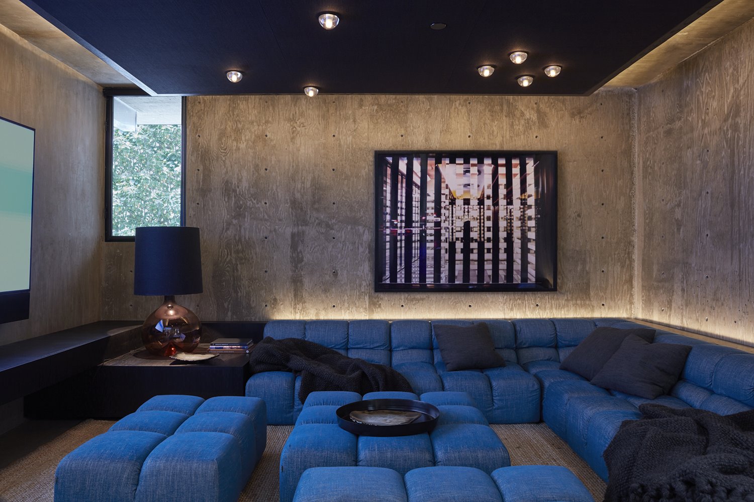 Sala de cine con sofás y poufs en azul, paredes de hormigón, luminaria de sobremesa con pantalla negra y gra pantalla de televisión