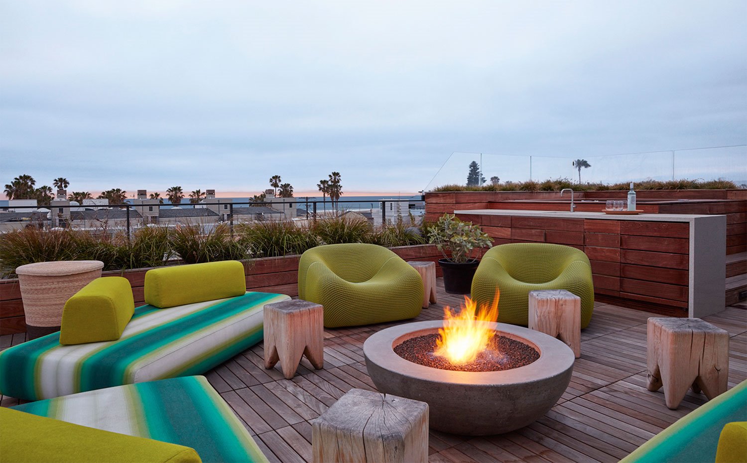 Terraza con fuego central, taburetes de madera, butacas en verde pistacho y cocina exterior revestida de madera