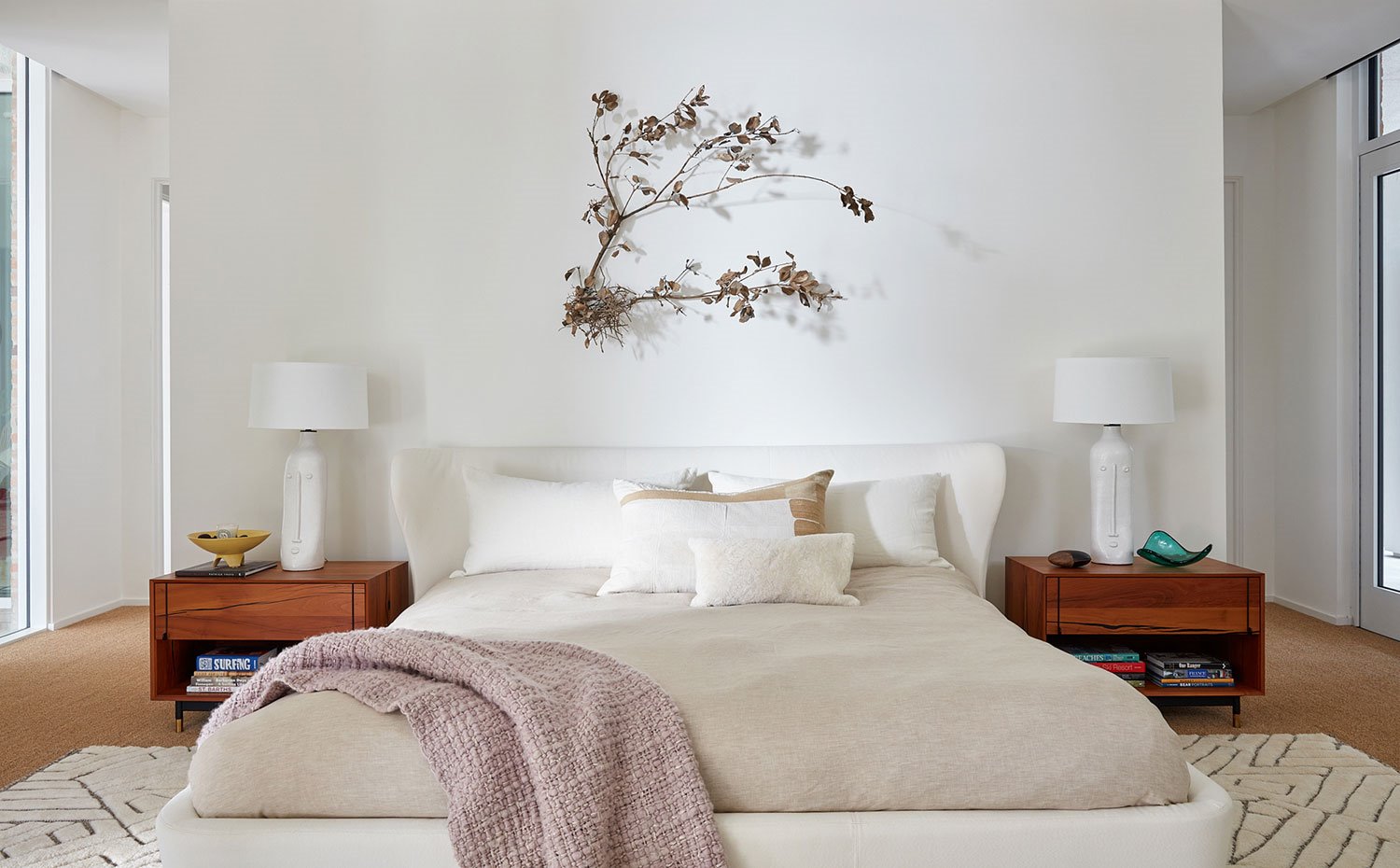 Cama principal con estructura en blanco, mesillas de madera, luminarias de sobremesa en balnco, detalle floral sobre cabecero