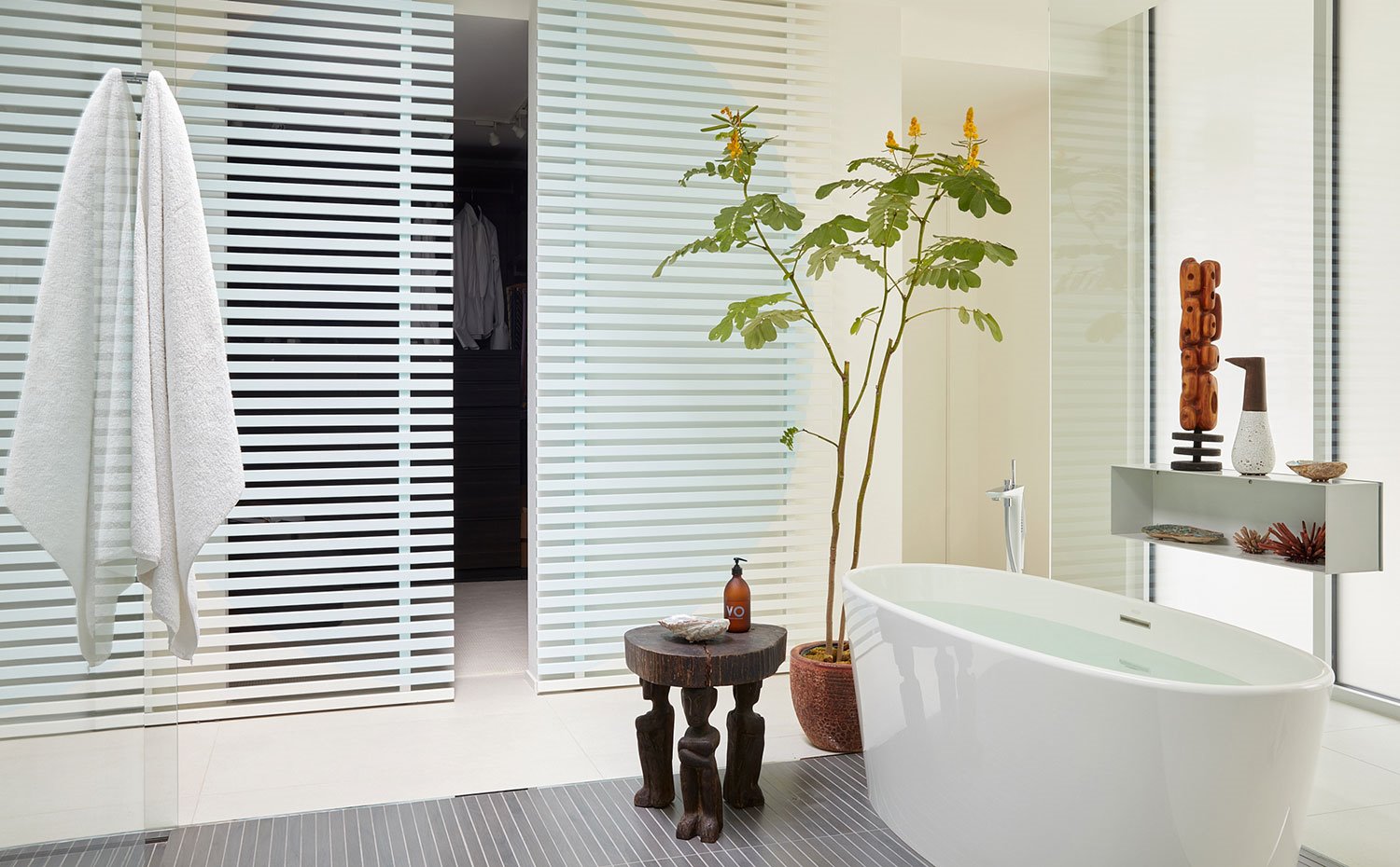 Cuarto de baño con bañera exenta, taburete de madera, cerramientos de cristal y puerta corredera hacia vestidor