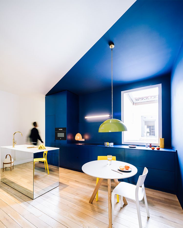 Cocina con mobiliario y pared en azul, techo inclinado, isla revestida de espajo, mesa circular para office