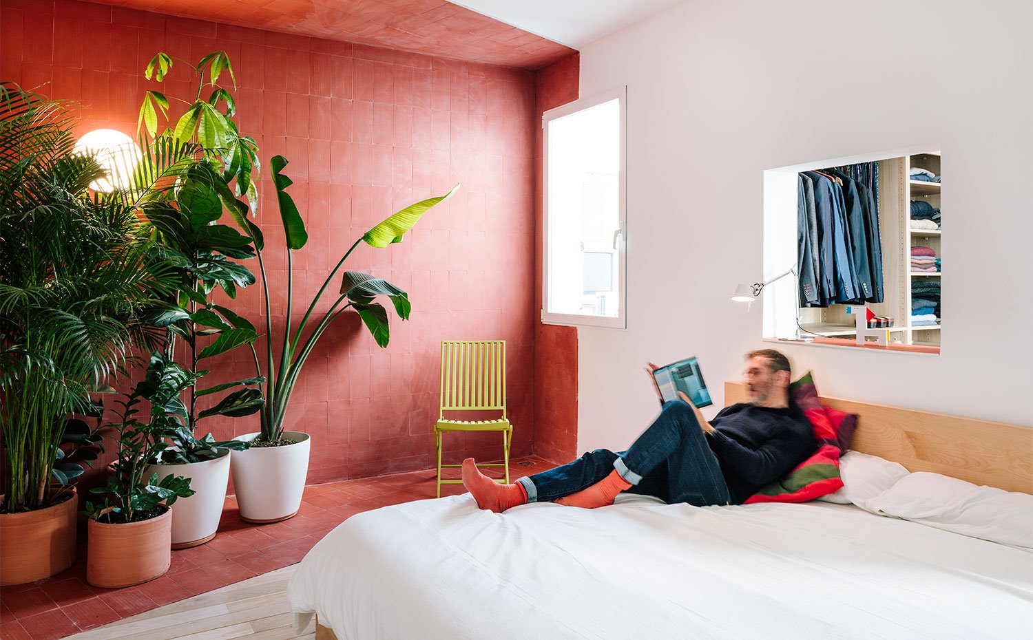 Dormitorio con hueco en pared sobre cabecero, pared pintada en tonos teja, grandes macetas con plantas, silla de madera en tonos verdes