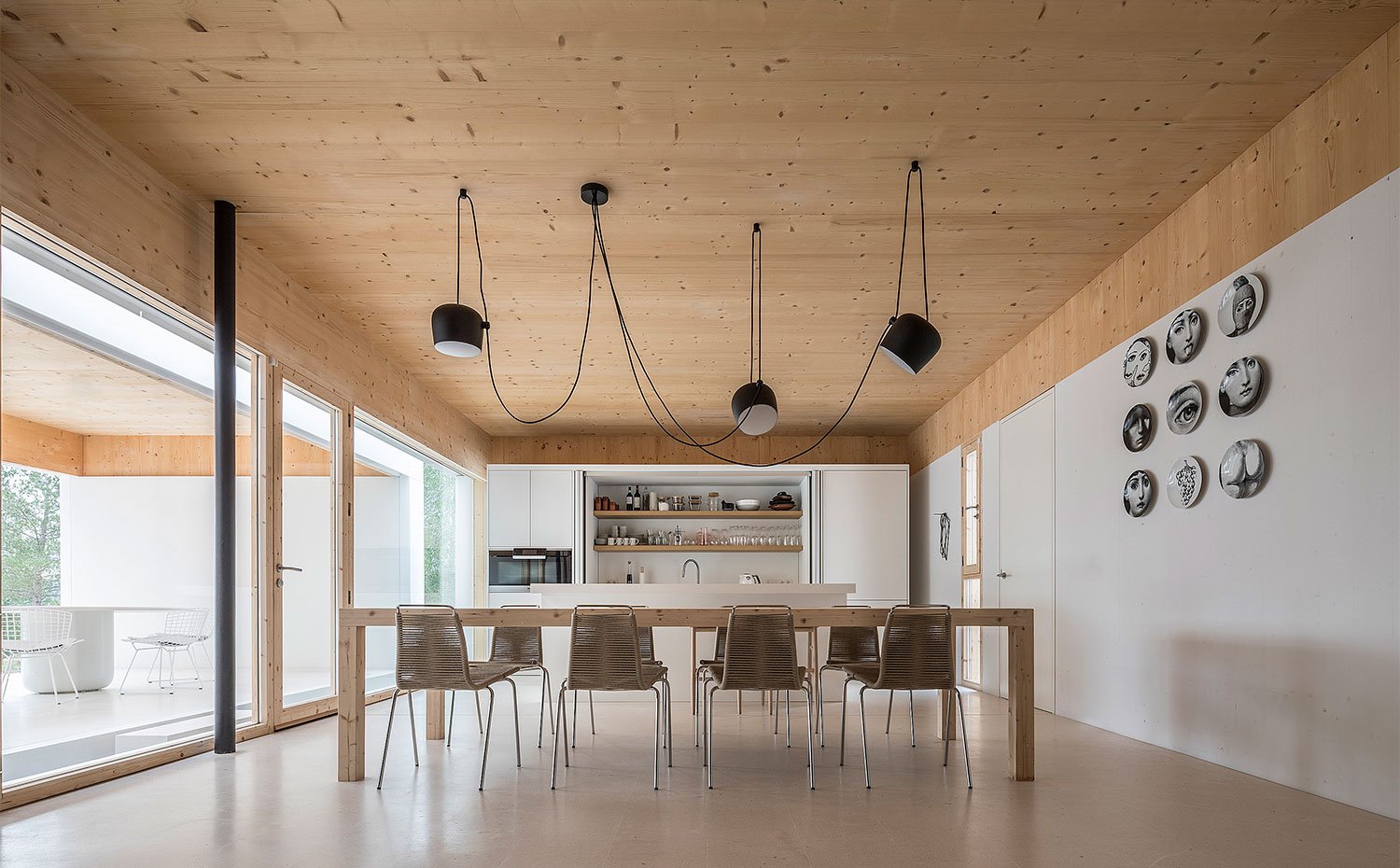 Comedor con amplia mesa de madera, luminarias suspendidas en negro, cocina abierta