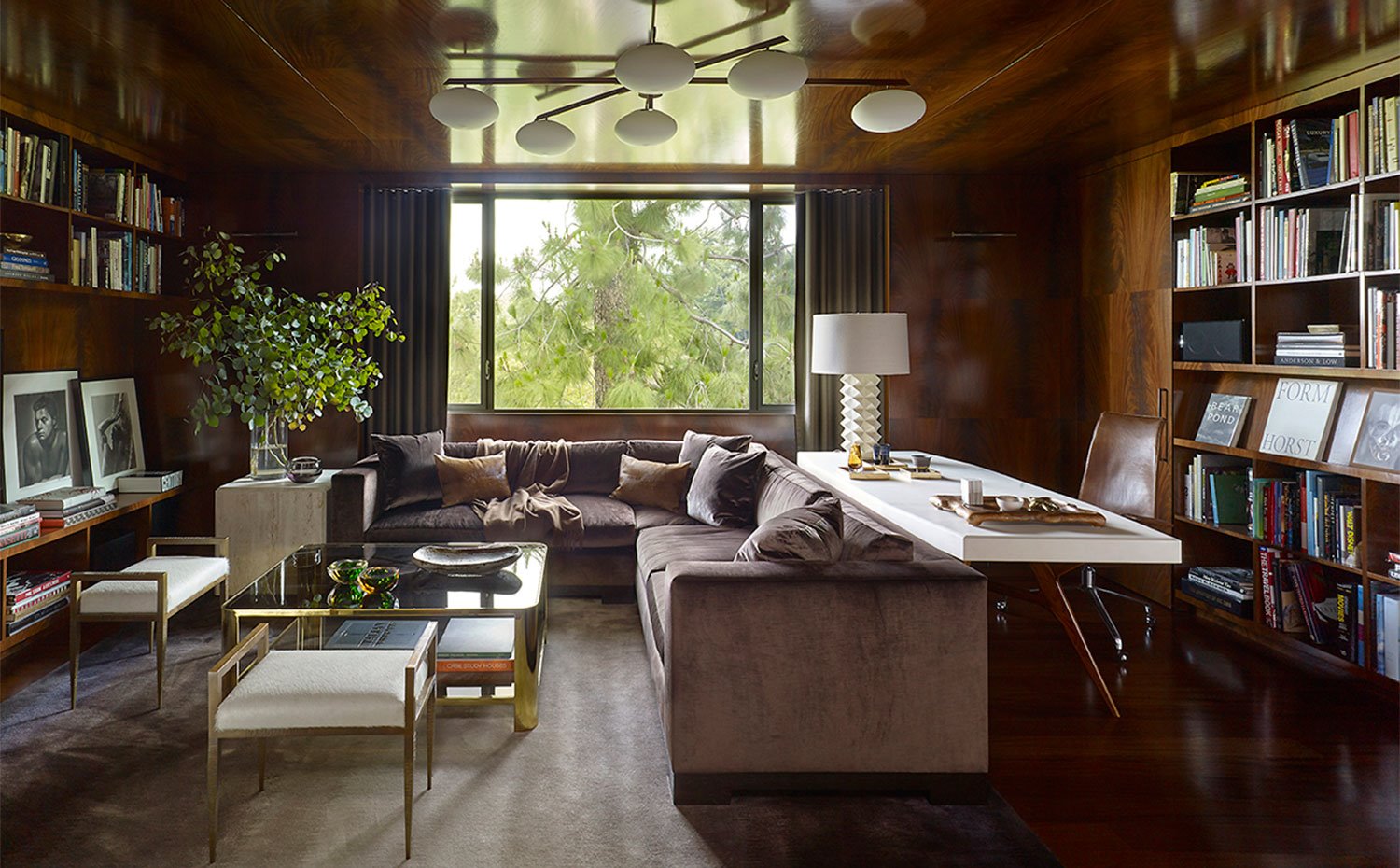 Biblioteca revestida de madera, con estanterías hechas a medida, sofa en L en tonos burdeos, mesa de centro en cristal, zona de trabajo con butaca tapizada en piel marrón