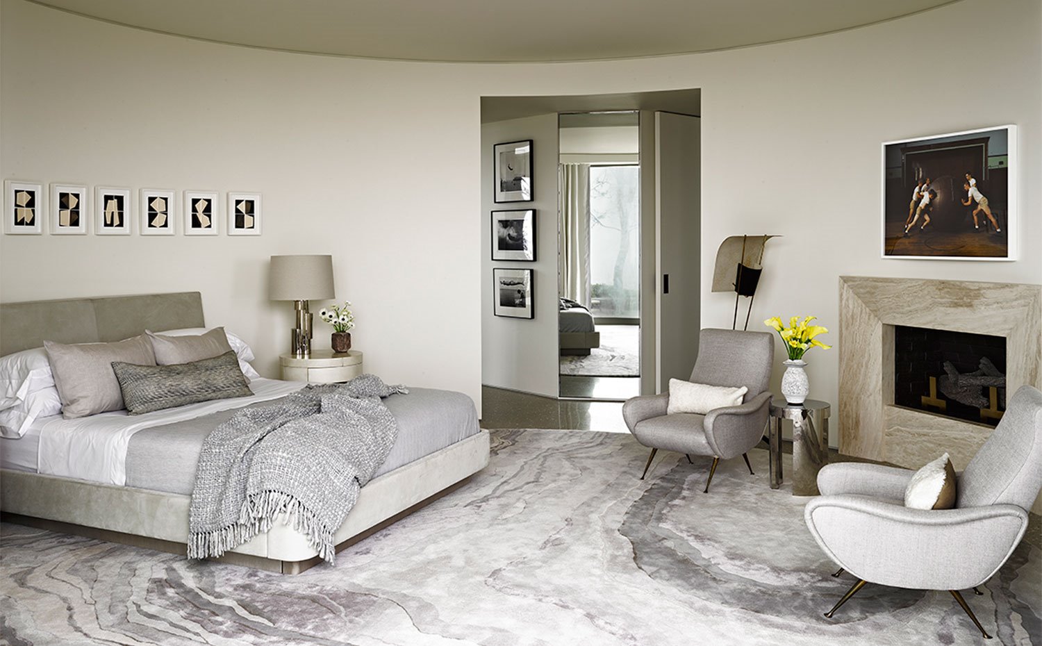 Dormitorio con alfombra de gran tamaño en tonos grisáceos, butacas grises, chimenea revestida de mármol y cama tapizada en tejido con textura aterciopelada.
