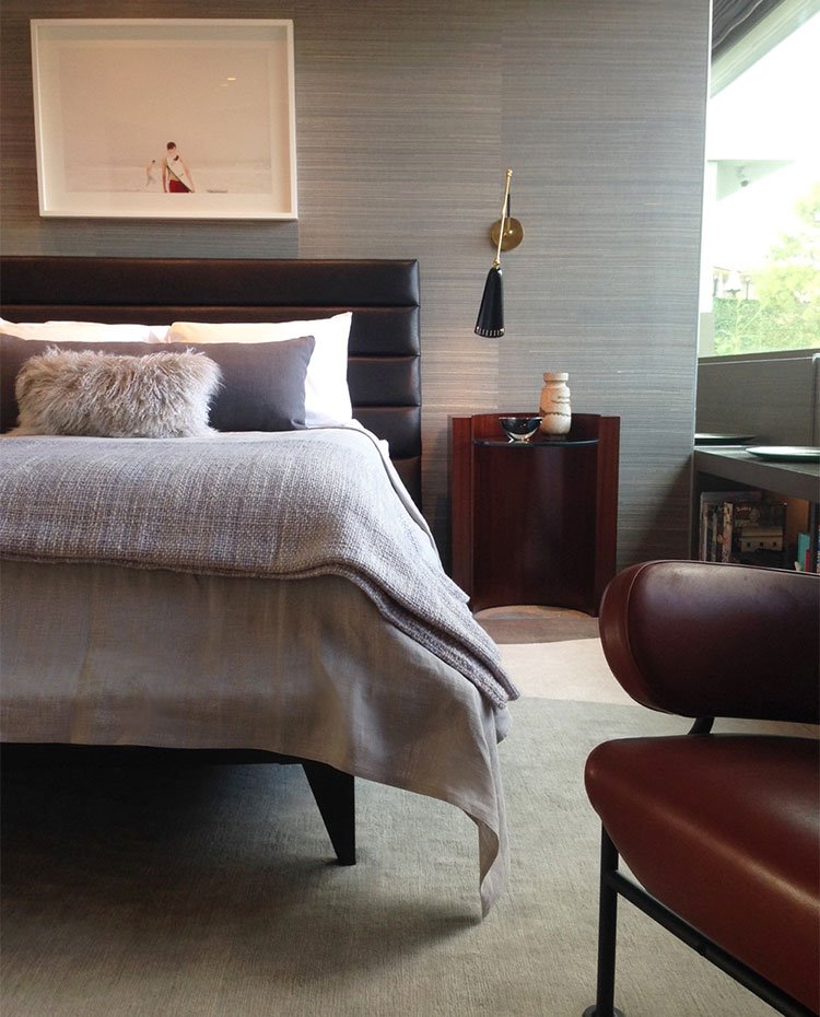 Dormitorio con cabercero revestido de piel marrón, papel pintado en pared en tonos grisáceos, butaca de piel marrón, mesita de noche en madera, aplique con acabdos dorados