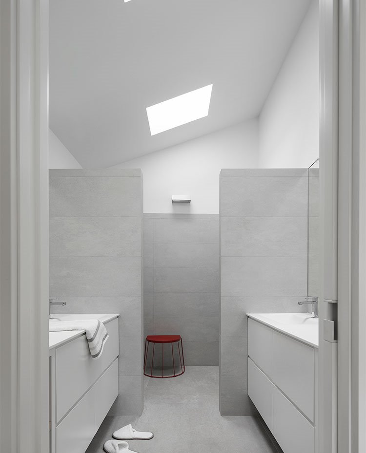 Cuarto de baño revestido en gris, con mobiliario en blanco y lucernarios en el techo