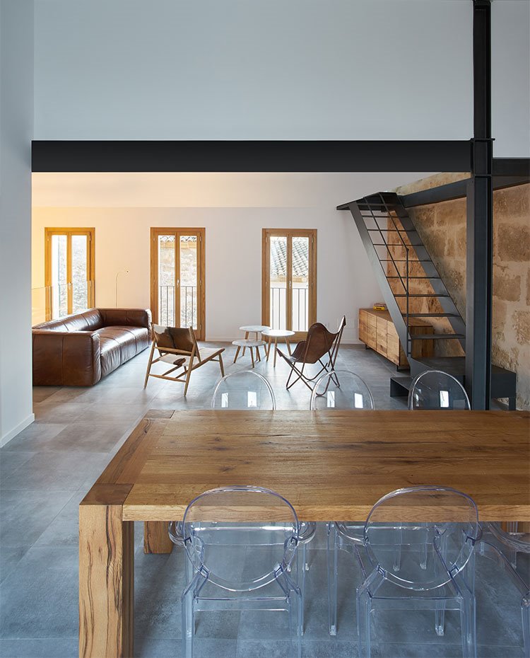 Mesa de comedor de madera y sillas transparentes, con salón de fondo y escaleras de hierro
