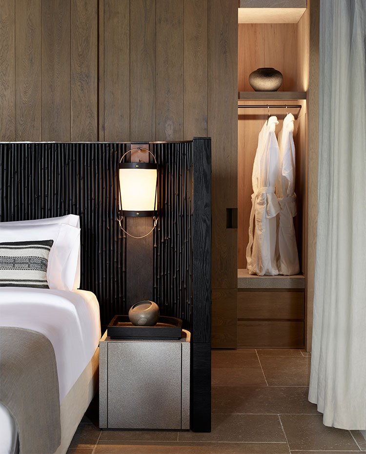 Dormitorio con cabecero oscuro, con suelo porcelánico y armario de madera oscura con coritnas en crudo