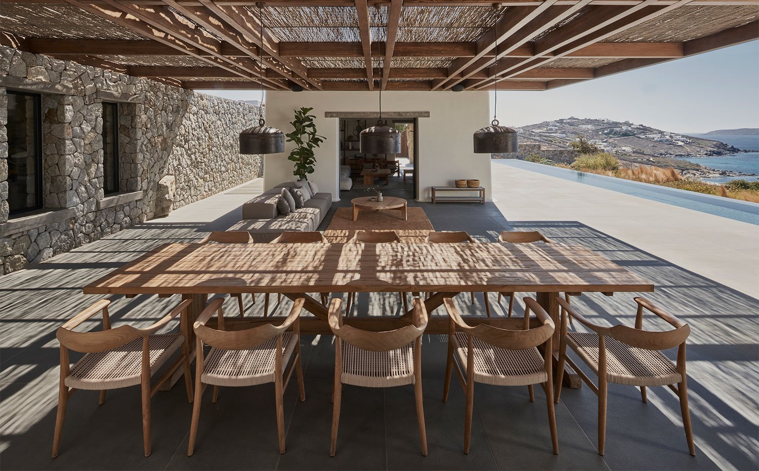 Comedor exterior con amplia mesa y sillas de madera con zona de estar en segundo término bajo porche