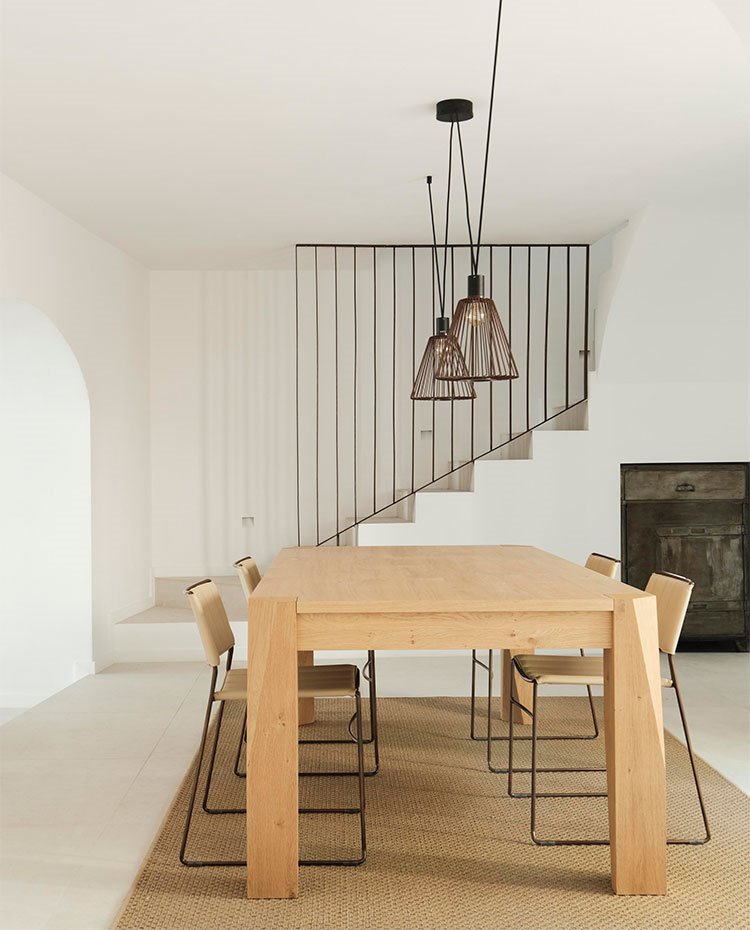 Comedor con mesa de madera y sillas con estructura metálica, luminarias suspendidas en negro, escaleras al fondo