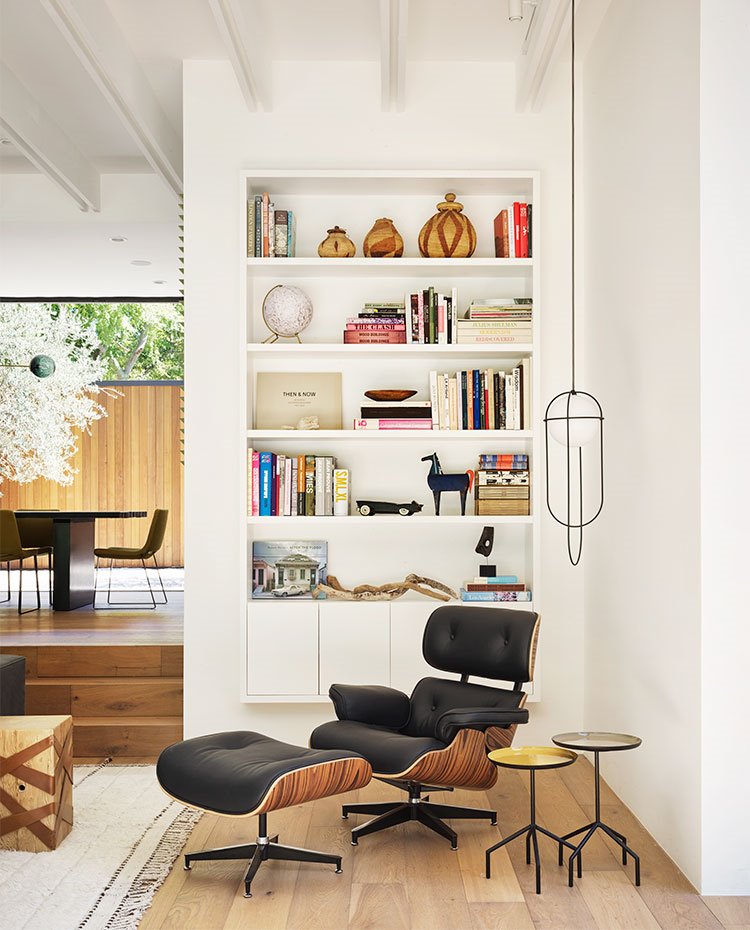 Rincón de lectura en salón con butaca y pouf en madera y piel, luminaria suspendida y librería en blanco suspendida en pared