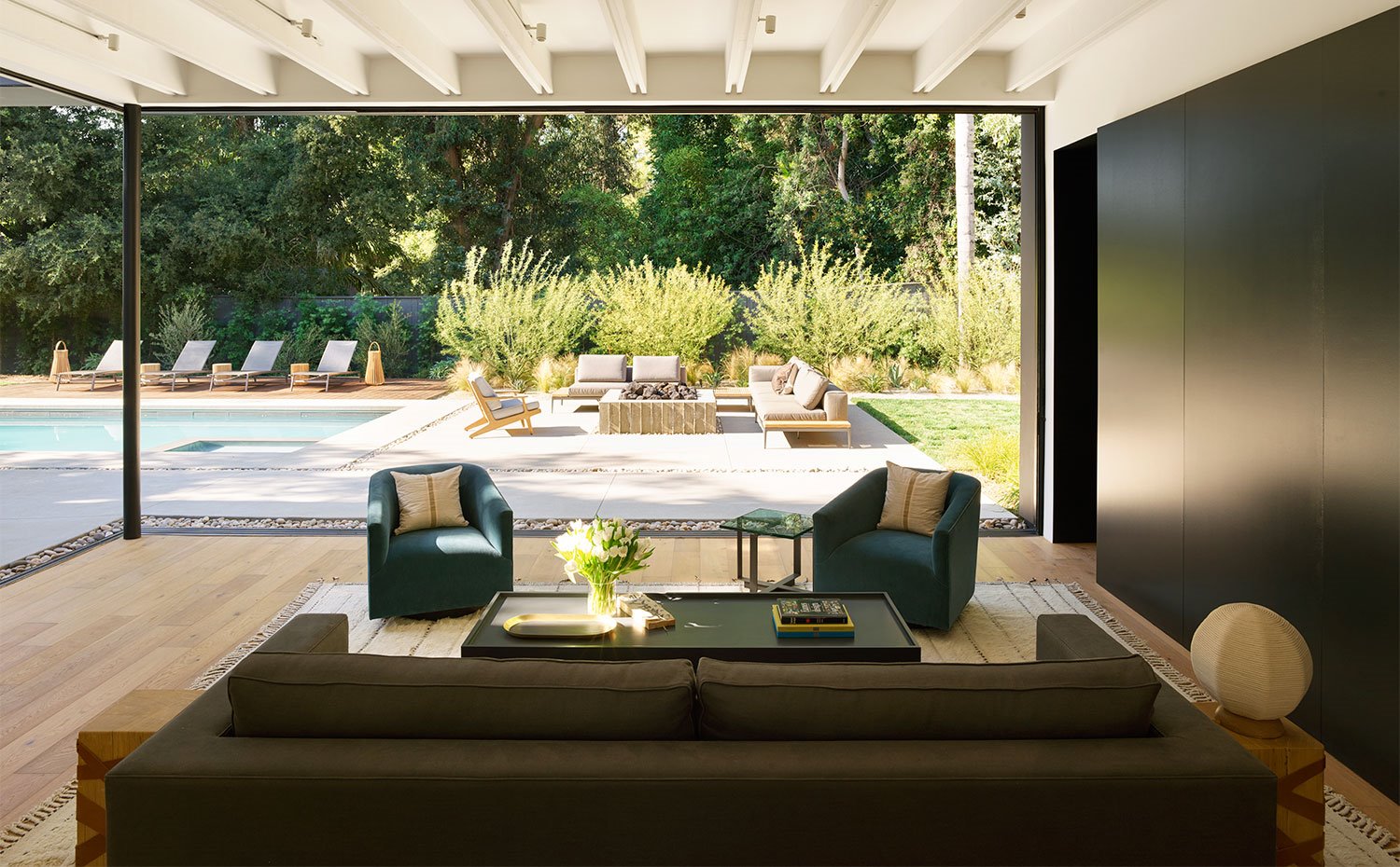 Salón con sofá y butacas en tonos oscuros, suelo de madera y acceso abierto hacia terraza y piscina