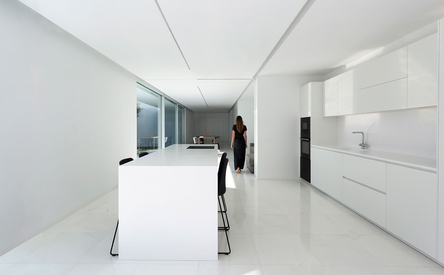 Cocina con amplia isla central, todo en blanco con taburetes y columna de electrodomésticos en negro