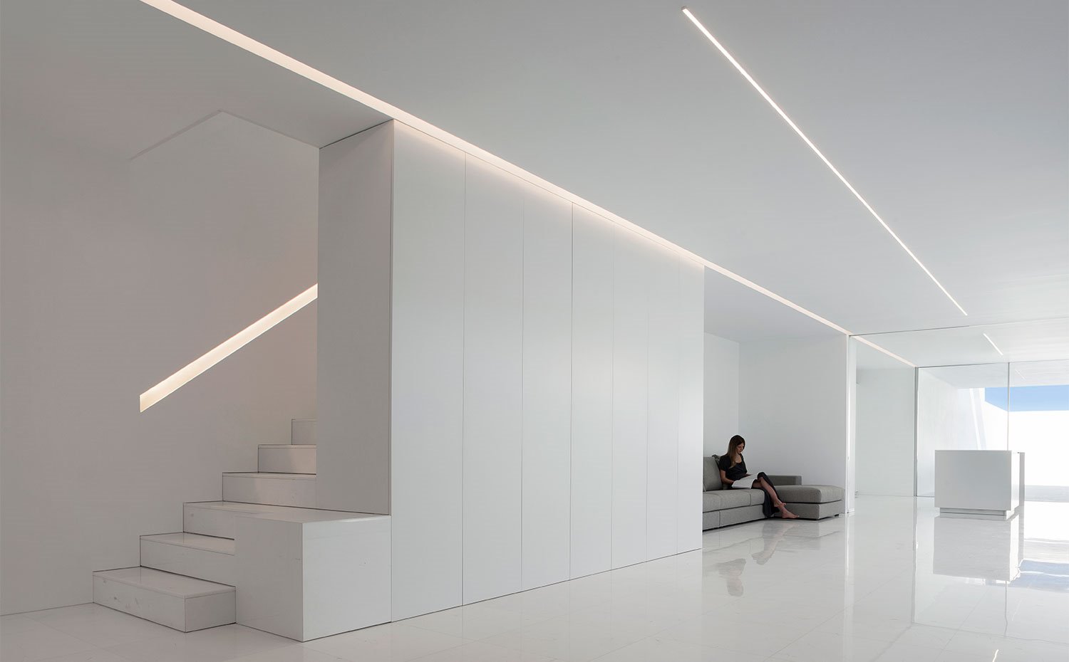 Zona de estar con iluminación lineal y escaleras, todo en blanco