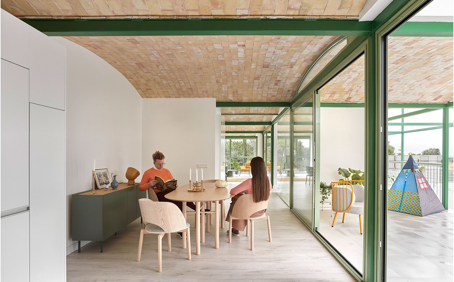 Comedor interior abierto a terraza exterior con mesa y sillas en madera y módulo de almacenaje en verde 
