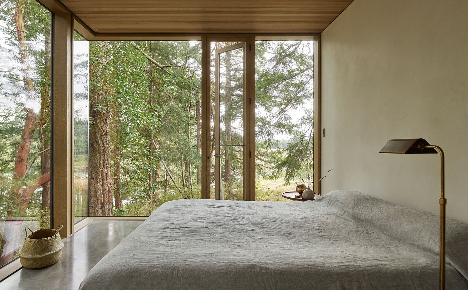 Dormitorio con techo y perfilería en madera, luminaria de pie en latón, cesto de mimbre y cerramiento acristalado