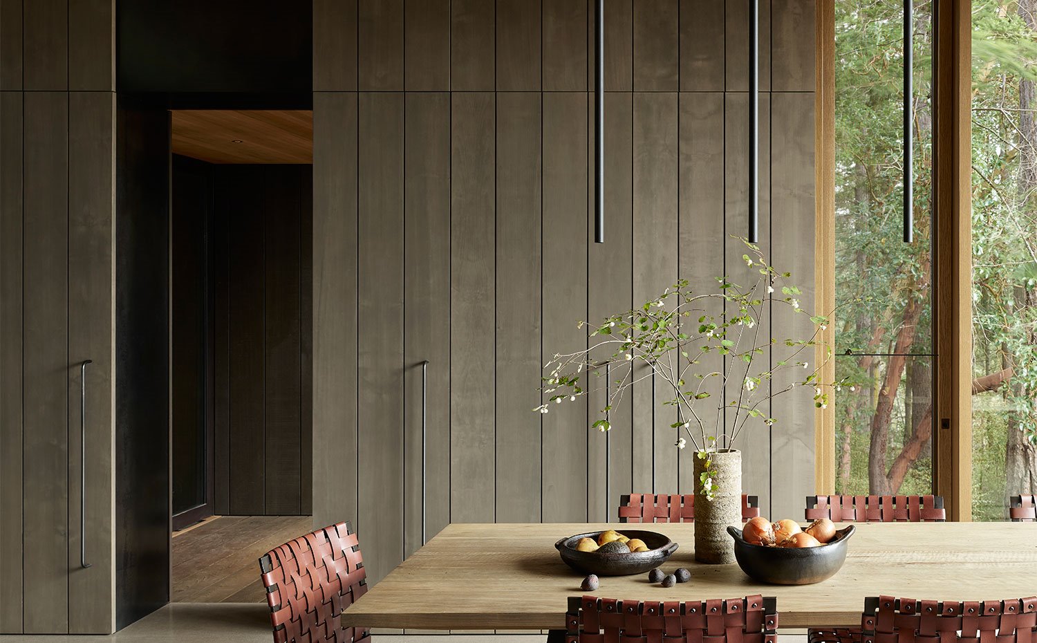Mesa de comedor en madera y sillas con trenzado de piel, armarios alistonados de madera oscura con largos tiradores metálicos