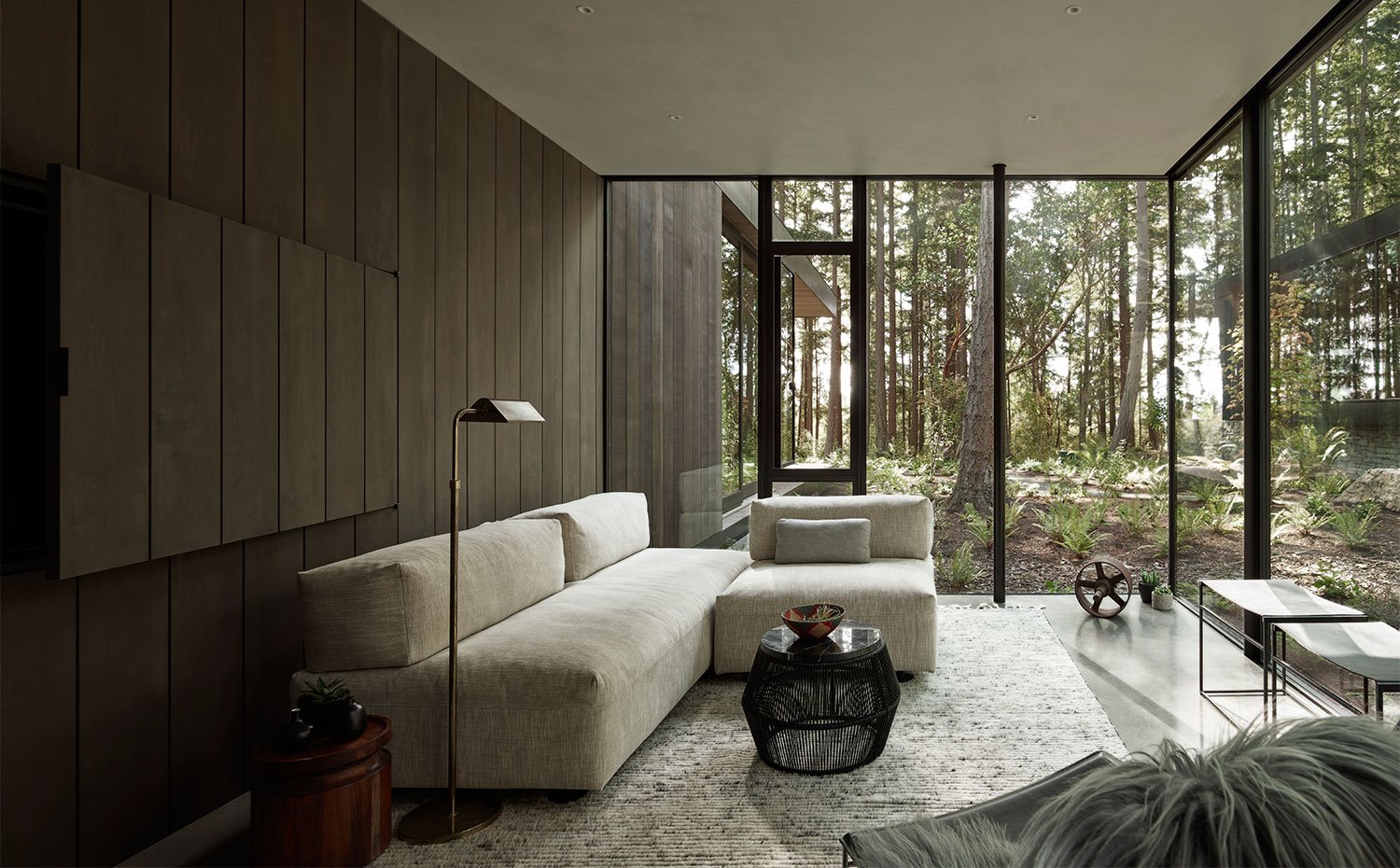 Sala de estar con revestimiento en madera oscura, sofá en tonos ocres y amplios cerramientos de cristal