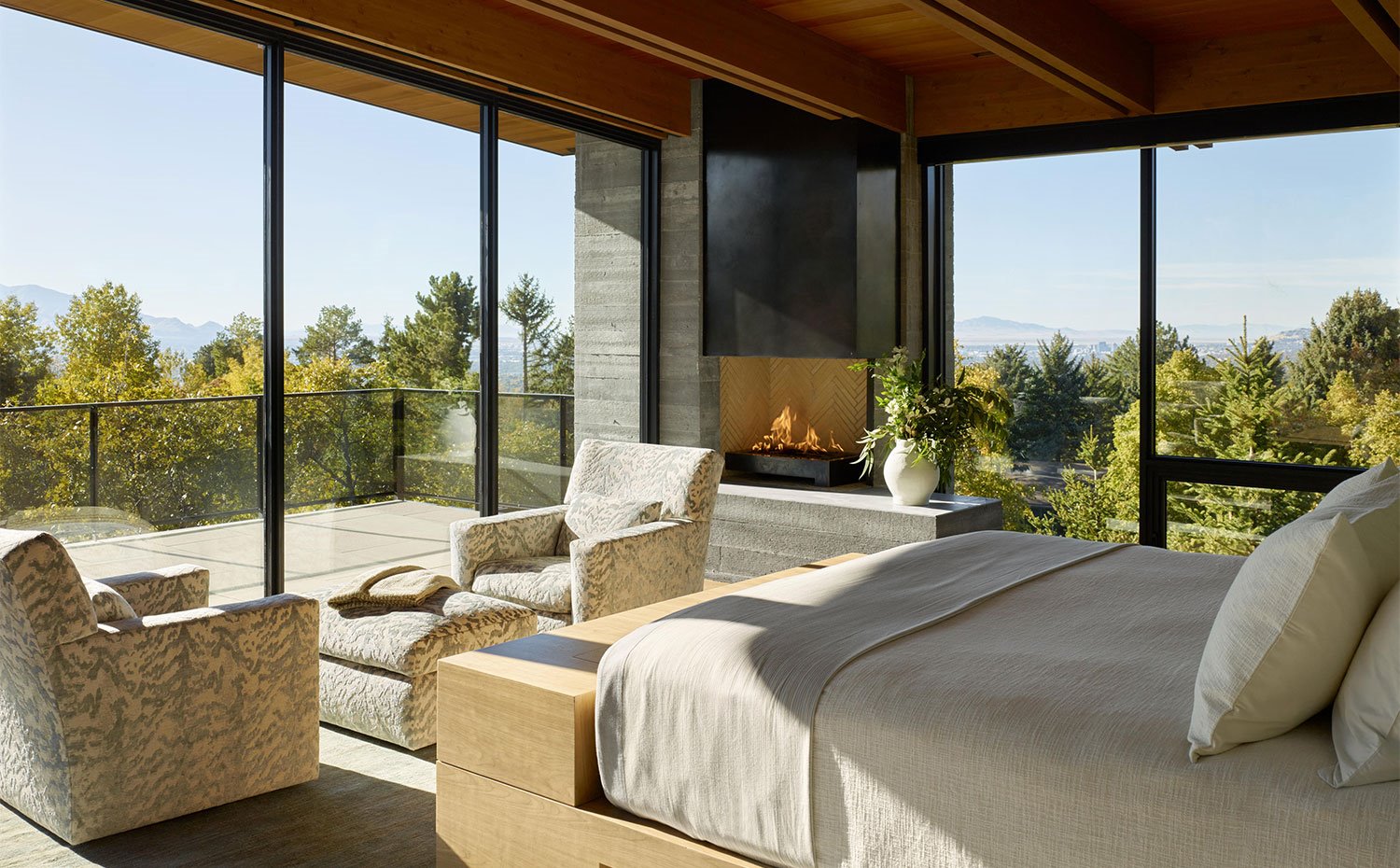 Dormitorio con balcón exterior, estructura de cama en madera, chimenea en la esquina y cómodos sillones con reposapiés a juego