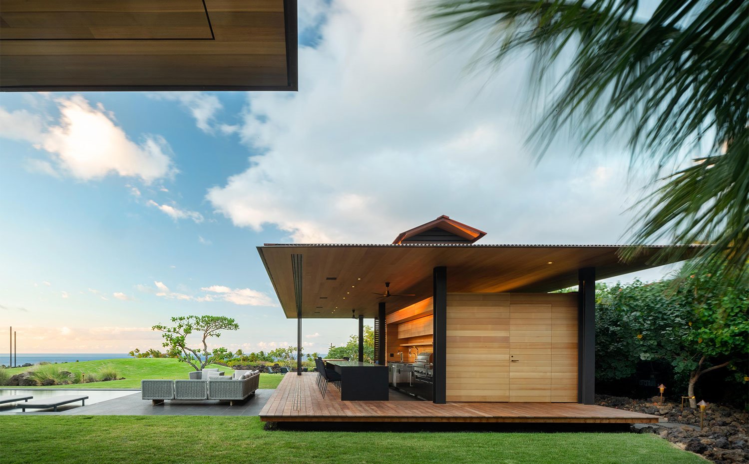 Módulo con cocina exterior con barbacoa e isla con taburetes, junto a mobiliario exterior situado al borde de la piscina