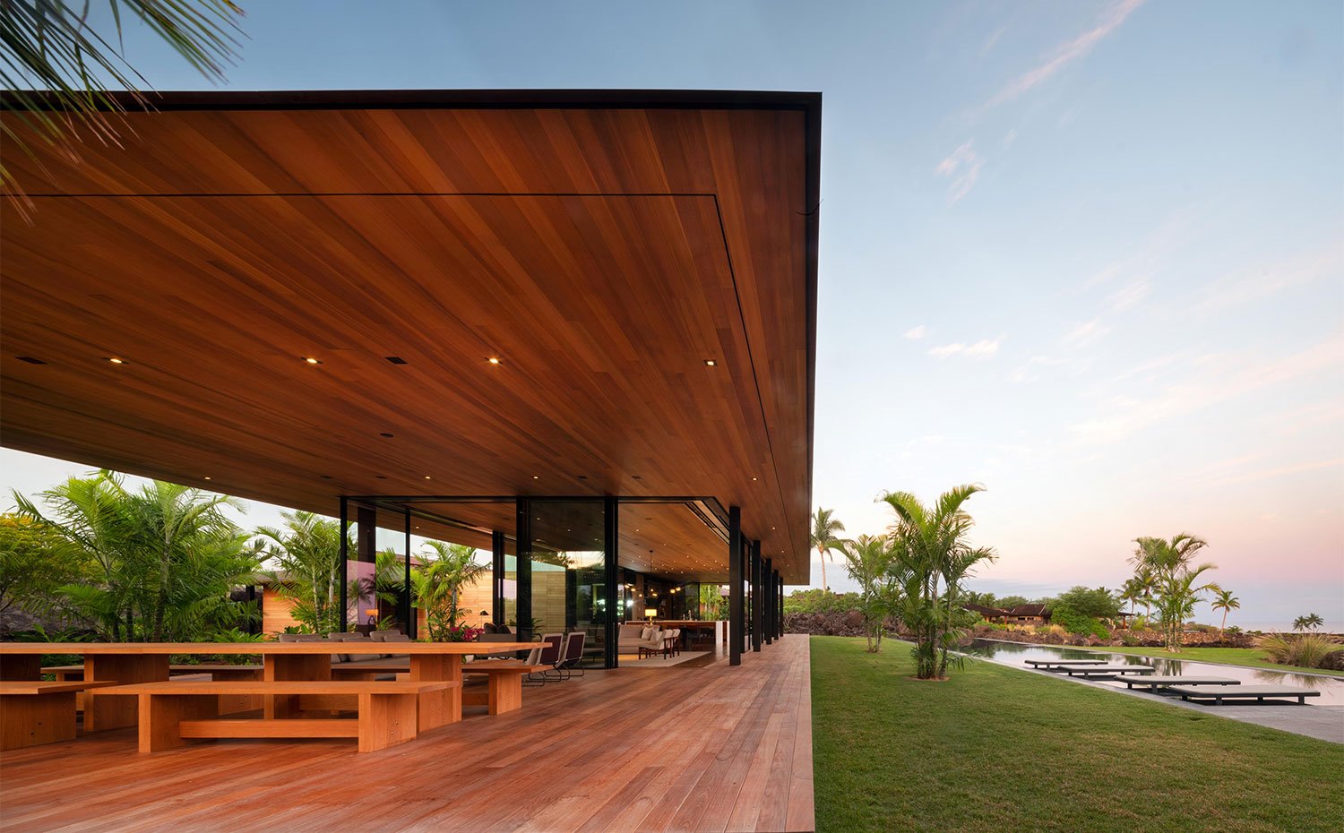 Vista del comedor exterior sobre plataforma de madera cubierta, junto a zona ajardinada y piscina