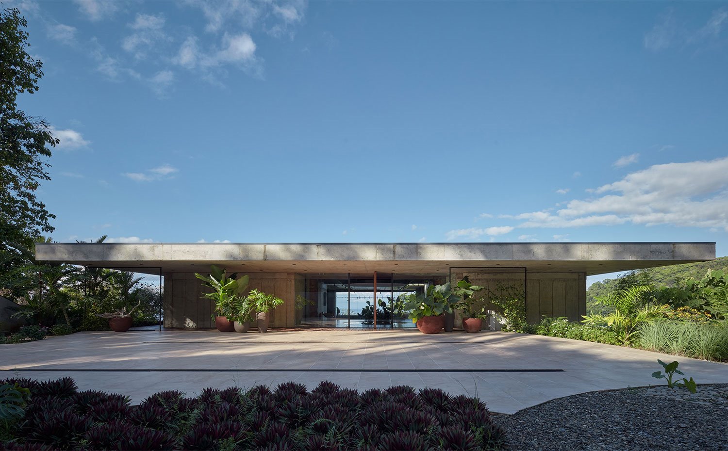 Frontal de la casa con voladizo de hormigón rodeada de vegetación y plantas tropicales en grandes macetas