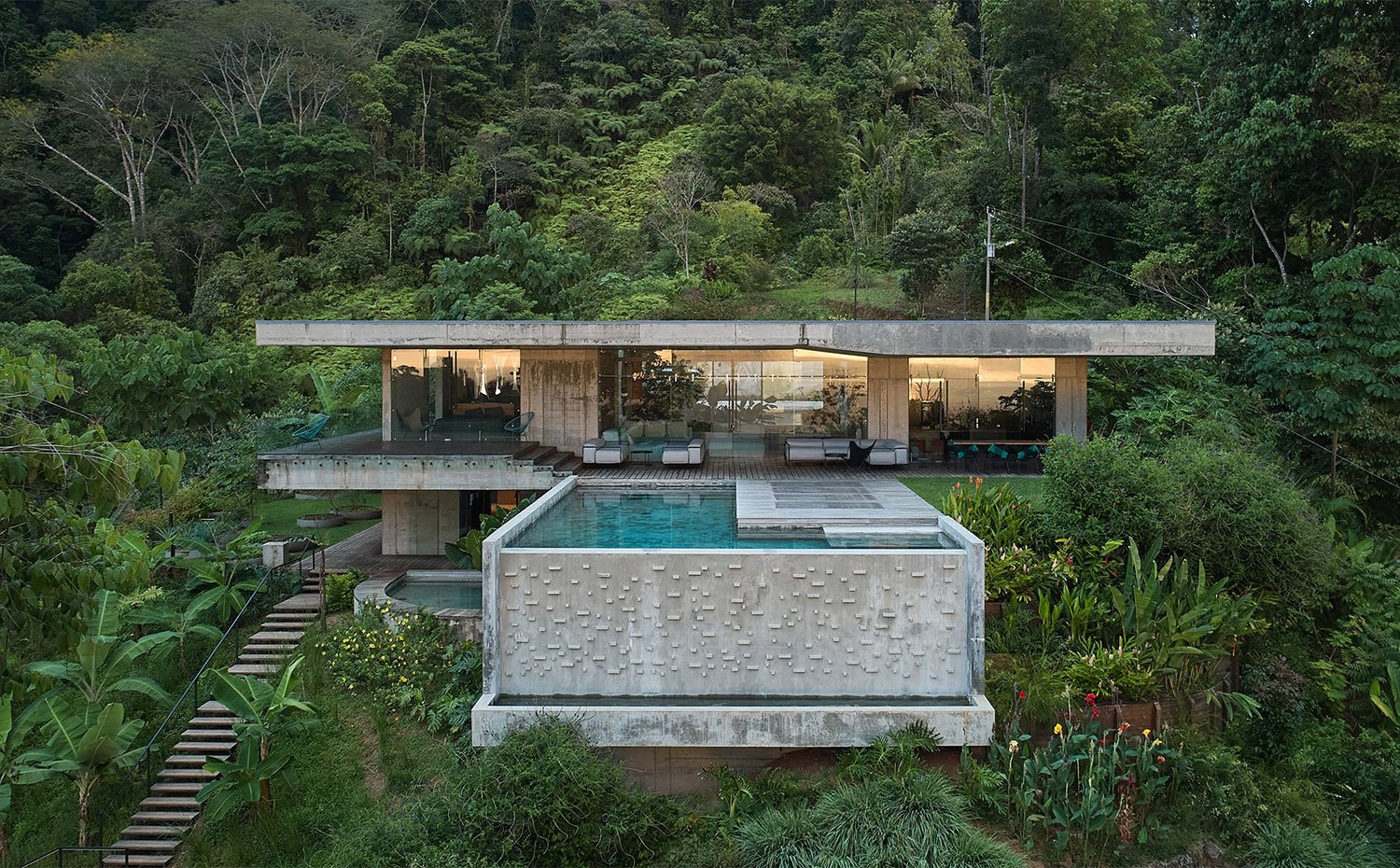 Vista general de la vivienda con gran piscina junto a terraza y escaleras de madera entre densa vegetación