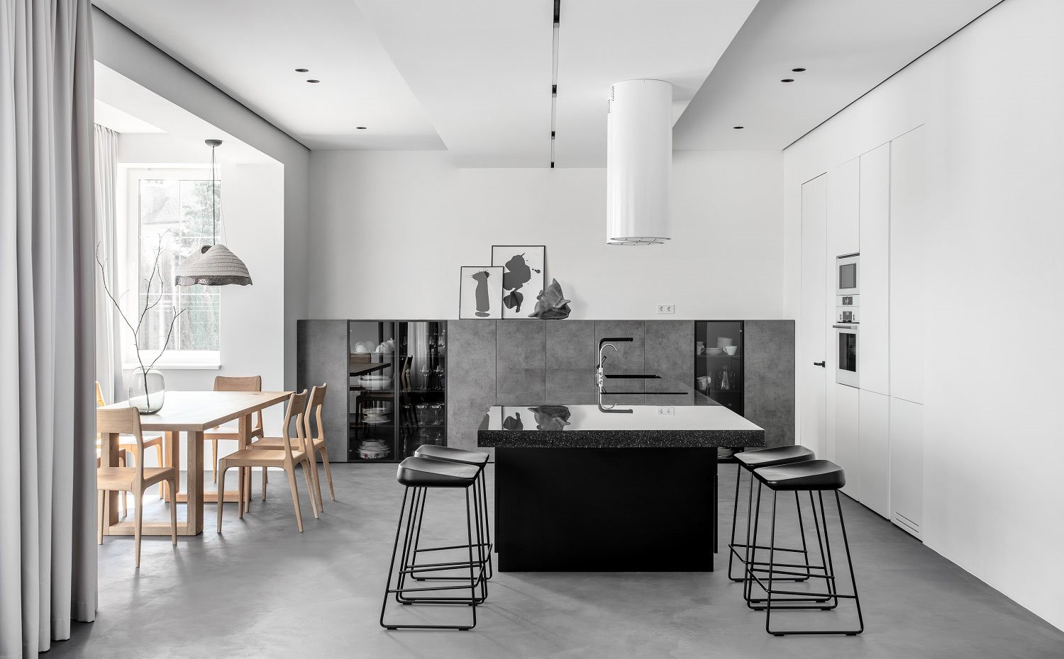 Cocina y comedor en un solo ambiente, con mobiliario en blanco, gris y negro y campanas estractoras cilíndricas en blanco