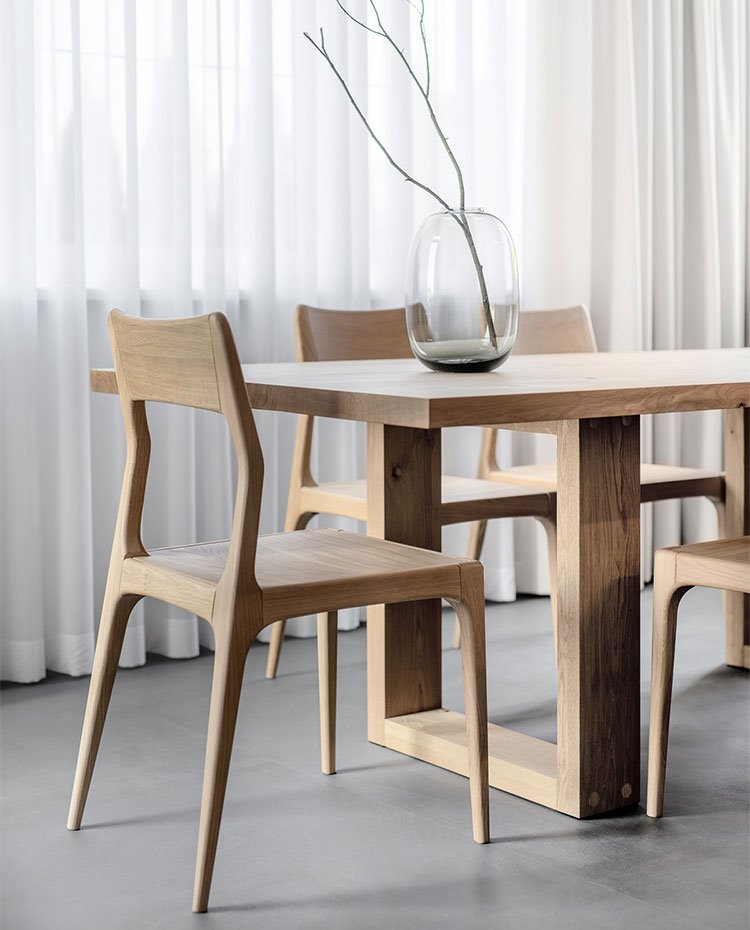 Detalle sillas y mesa de comedor en madera, con jarrón de cristal y cortinas blancas al fondo