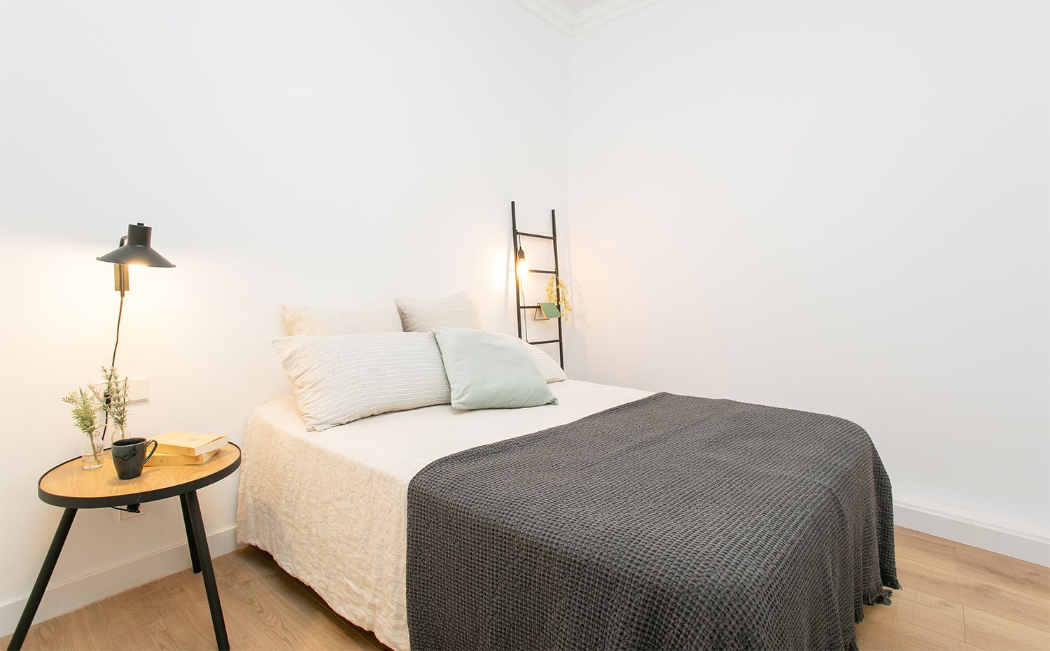Dormitorio con suelo de madera, ropa de cama en blanco y gris, mesilla de noche con aplique en negro
