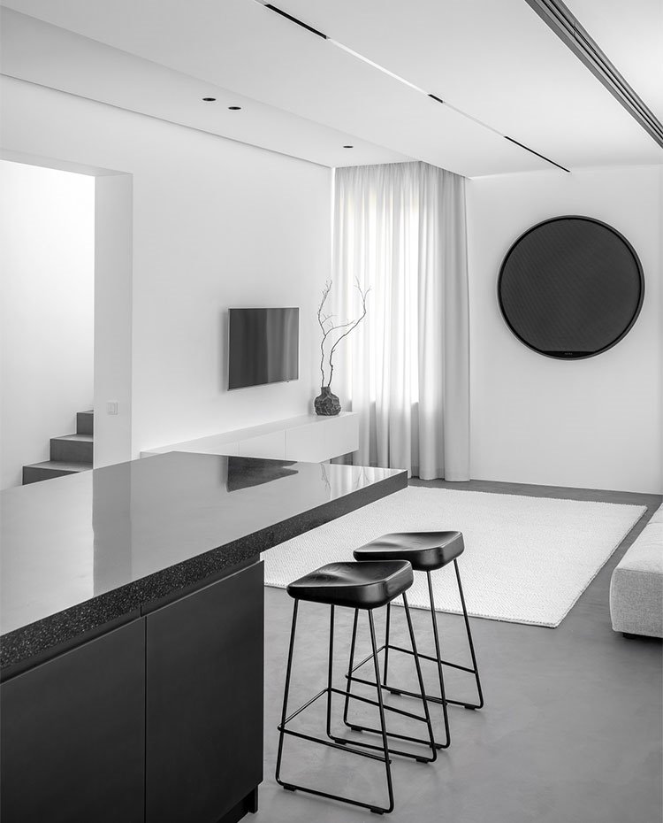 Taburetes del office de cocina, en negro, junto a salón con alfombra blanca sobre suelo en gris oscuro y mobiliario del salón en blanco