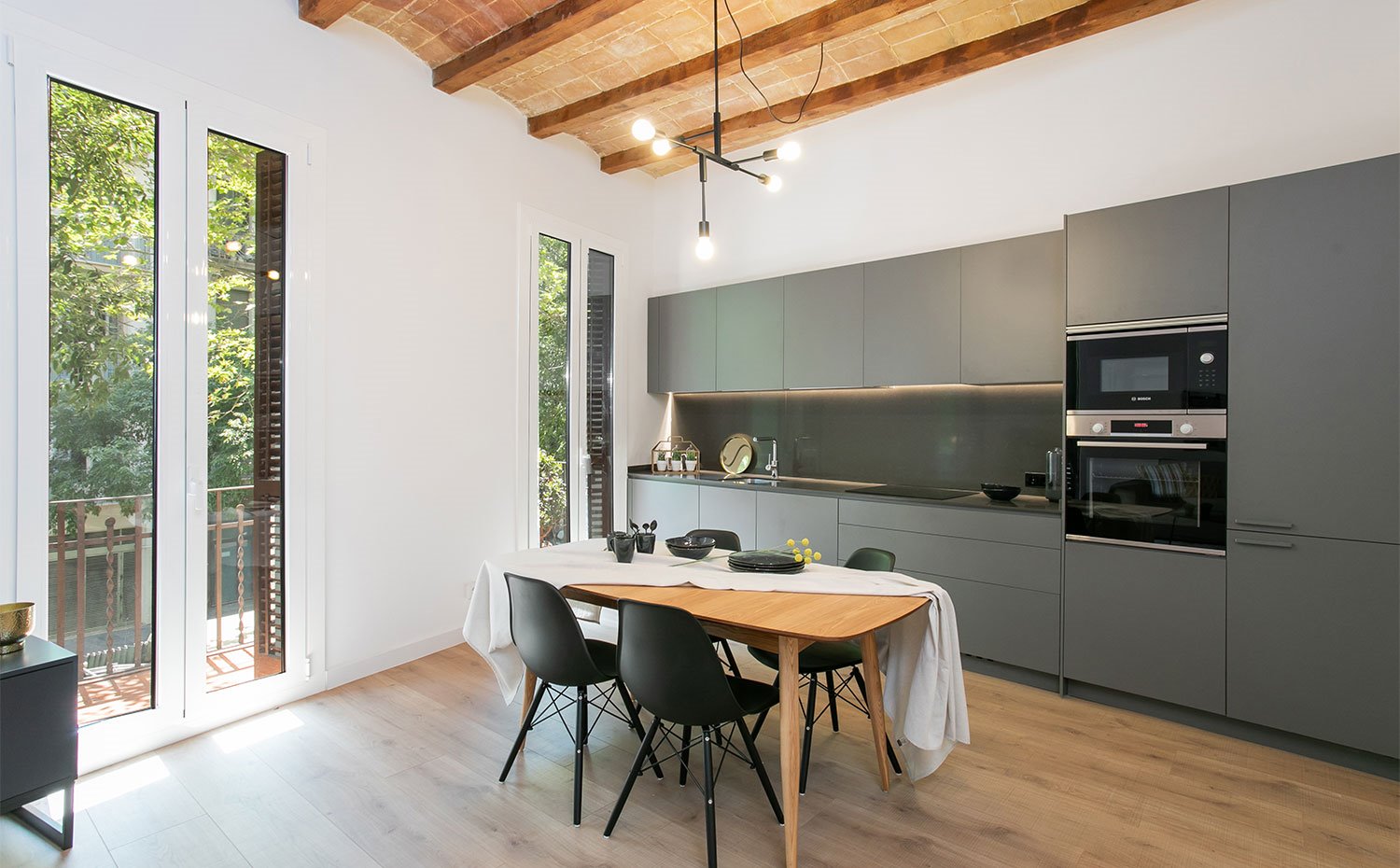Techo abovedado con vigas de madera, cocina en gris abierta a comedor y salón, mesa de comedor de madera y sillas en negro